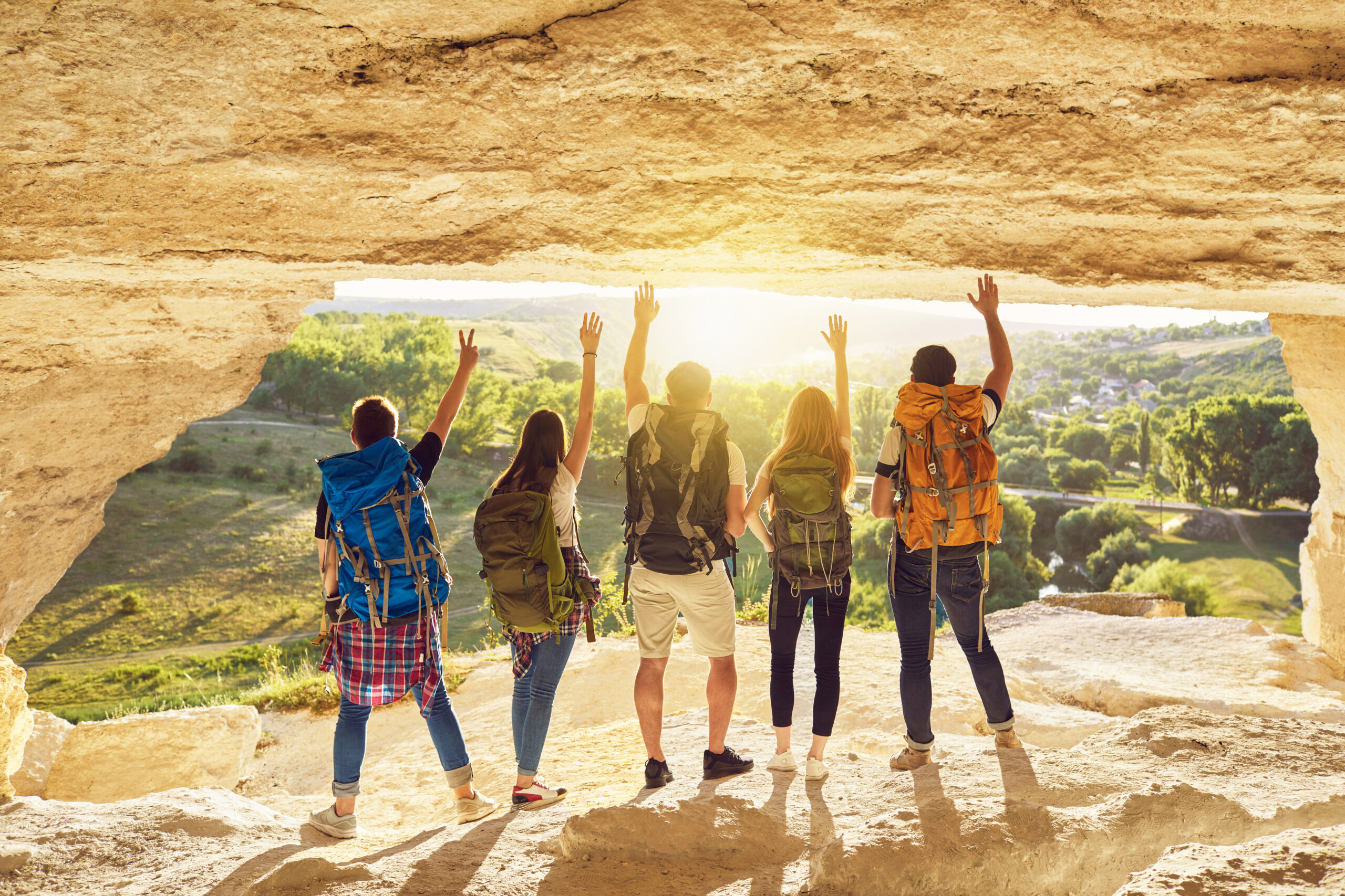 Great achievement. Back view of group of tourists with backpacks standing arms raised at cave entrance on mountain top admiring magnificent nature. Young hikers traveling during summer vacation